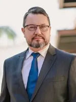Alexandre Strohschoen em traje formal com terno escuro e gravata azul, posando ao ar livre em ambiente