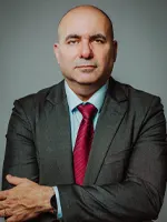 Ednei Aquino em traje formal, com expressão séria, vestindo terno escuro e gravata vermelha, posando de braços cruzados diante de fundo neutro.